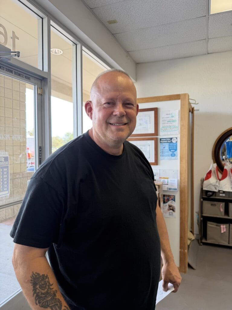 A man with a shaved head and tattoo on his forearm stands indoors by a door, wearing a black t-shirt and smiling at the camera.