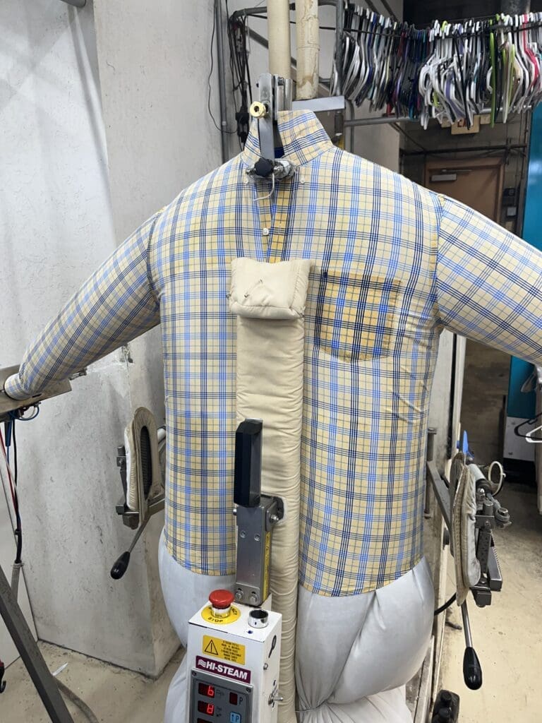 A plaid shirt is being steamed and pressed on an inflatable mannequin in a laundry or dry cleaning facility, with clothing hanging in the background.