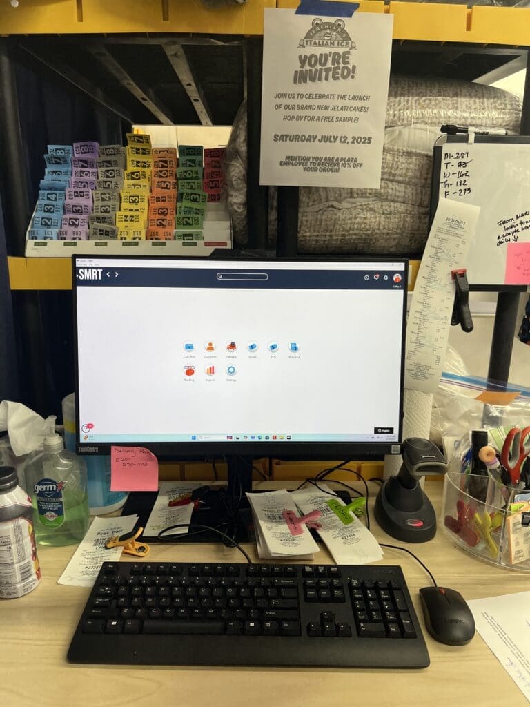 Desktop workstation with a monitor, keyboard, and various office supplies, including sticky notes and sanitizing wipes, set on a cluttered desk below a yellow shelf with colorful bins and a printed sign.