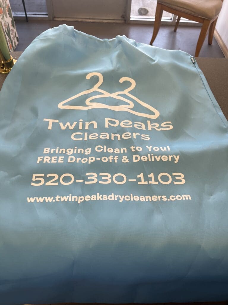 A blue Twin Peaks Cleaners laundry bag with white text showing the company name, logo, phone number, website, and a message about free drop-off and delivery service.