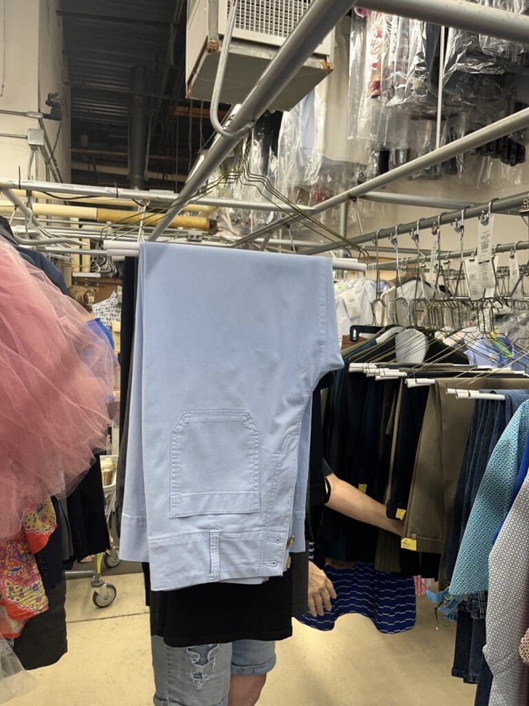 A person stands in a laundry or costume room with a pair of light blue pants hanging on a rack, obscuring their upper body and head.