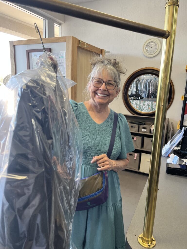 Woman in a teal dress smiles while picking up dry cleaning at a counter, holding a plastic-covered garment on a hanger.