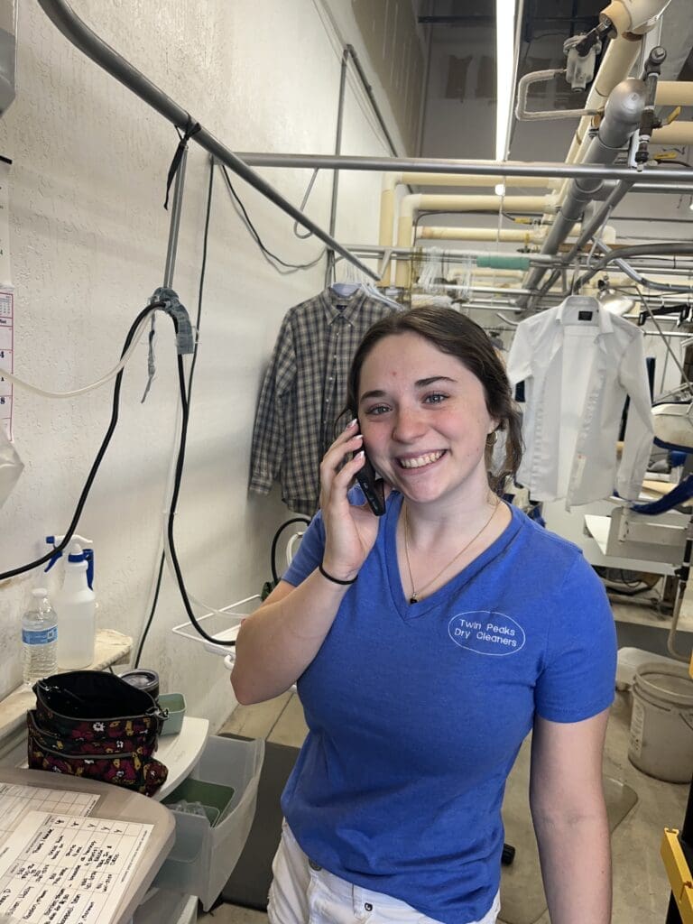 A young woman in a blue T-shirt stands in a laundry facility, smiling while talking on the phone. Laundry equipment and hanging garments are visible in the background.