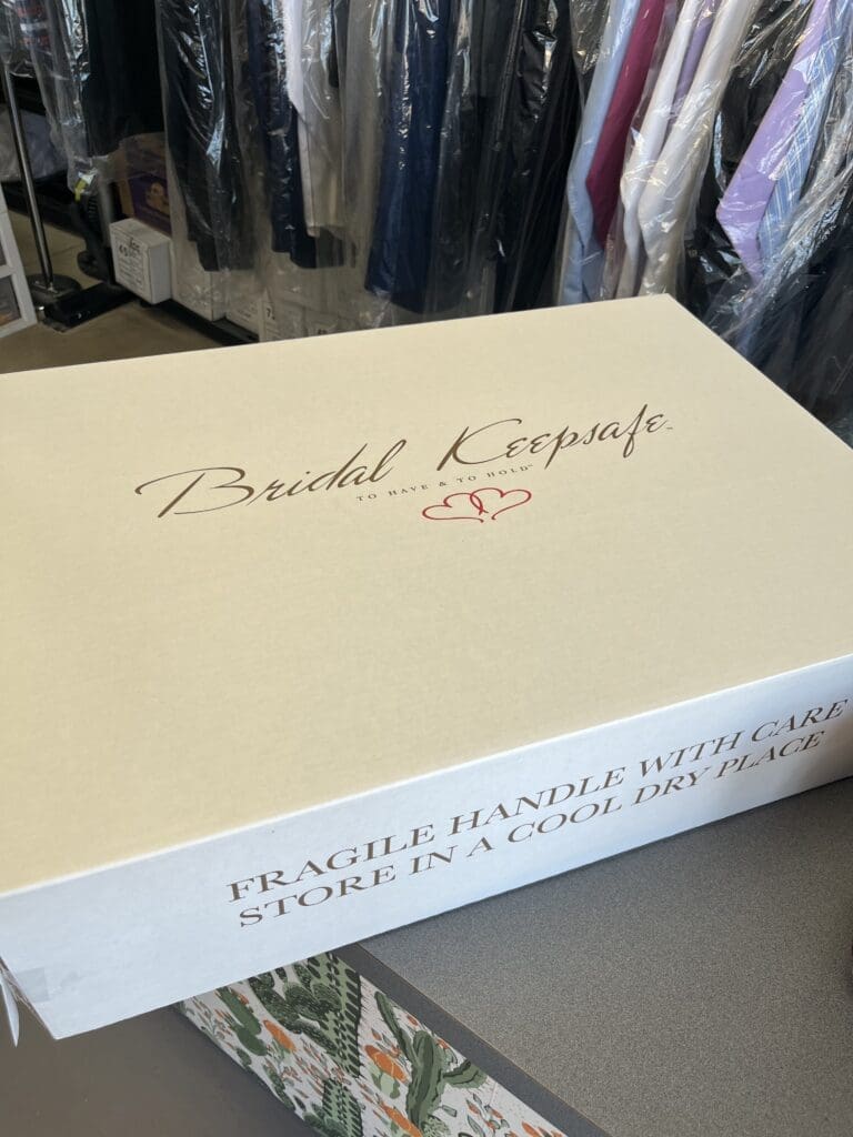 A white box labeled “Bridal Keepsafe” sits on a counter, with instructions to handle with care and store in a cool, dry place. Clothing racks are visible in the background.